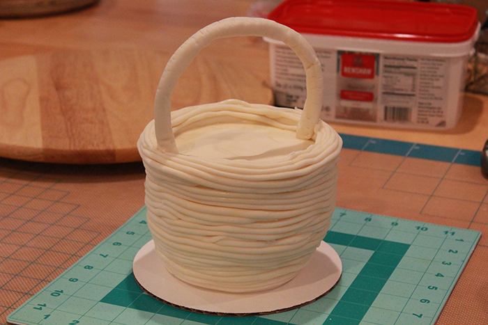 For the top tier, insert the basket handle, making sure to cover the exposed wire edges in a food safe product. Use ivory fondant and create a long snake roll. Wrap around the outside of the basket, working your way to the top of the basket edge. Use water to attach and then add some texture.