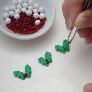 To create the holly for Santa’s hat, cut another sheet of wax paper to fit over the back of another baking sheet or other flat surface. Using the green piping icing with the Wilton #349 tip, pipe three connecting leaves. Pipe a second row of leaves creating a V shape. With the tweezers, place three small red nonpareils at the base of the holly leaves. Repeat until you have enough for each cookie. TIP: Royal icing transfers can be fragile so we always recommend making a few extra. Set the transfers aside and allow to completely dry (approximately 45 hours drying times vary depending on humidity). Leave transfers attached to the wax paper until you are ready to add them to the cookies.